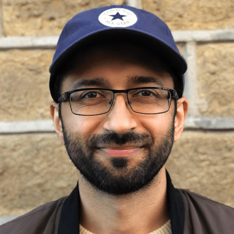 A tan-skinned young man with short hair and a neatly trimmed beard wearing glasses, a baseball cap and jacket smiles in front of a wall.