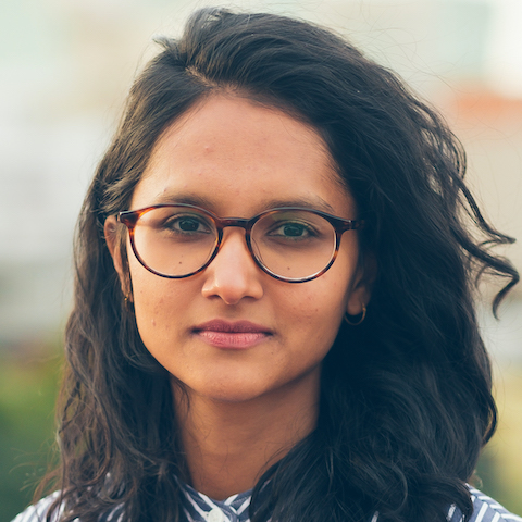 An outdoor portrait of a brown-skinned woman with shoulder-length black hair and glasses.