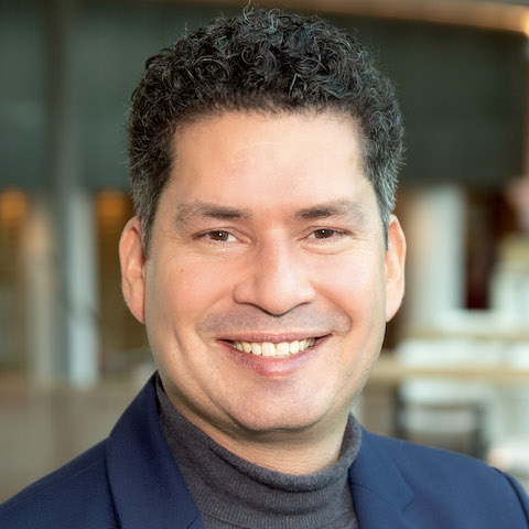 A professional portrait of a smiling white man in a turtleneck jumper and suit jacket with close-cut dark curly hair that's beginning to show signs of grey.