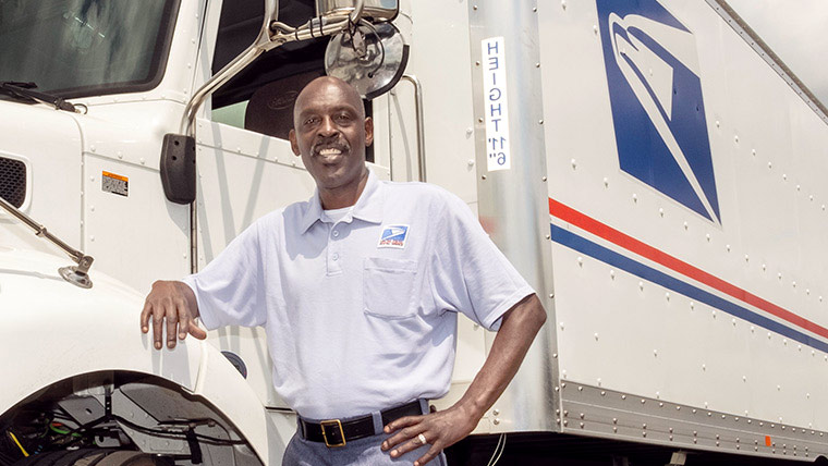 USPS employees standing in a group outside, in front of USPS delivery trucks.