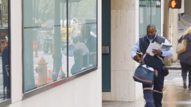 USPS Mail Carrier walking down a city sidewalk.