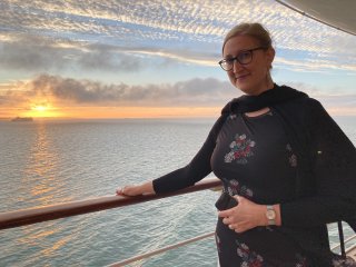 Jessica, dressed in an elegant outfit, standing on deck in front of a beautiful sunset over a calm sea.