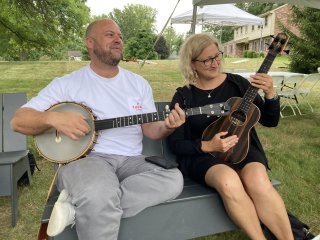 A man playing banjo and a woman playing bass ukulele on lawn furniture outdoors.