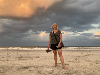 A woman stands holding her shoes on a sandy beach under a dramatic cloudy sky.