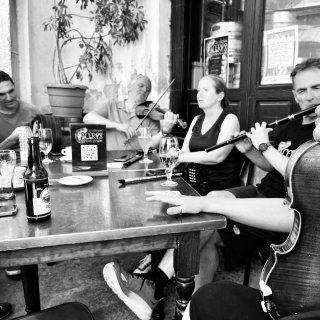 A group of musicians playing bodhrán, fiddle, concertina and flute playing at a table outside a bar.