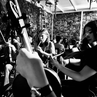 A hand holding a fiddle in front of a young woman playing fiddle and another woman playing tin whistle.