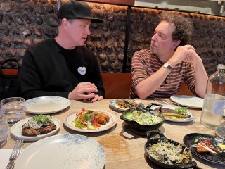 Two men turned towards one another at a tabled bedecked with lots of small plates of food.