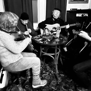 A circle of musicians: fiddles and tin whistle accompanying a singer playing guitar.