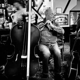 A hand is holding a fiddle and bow in front of a man playing flute in a pub.