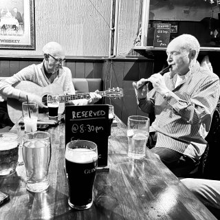 A man playing guitar accompanying a man playing tin whistle at a pub table bedecked with drinks.