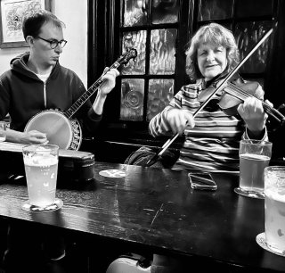 A man playing banjo alongside a woman playing fiddle at a pub table.