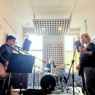 A young man on drums flanked by a man playing acoustic guitar on one side and a woman playing electric bass on the other.