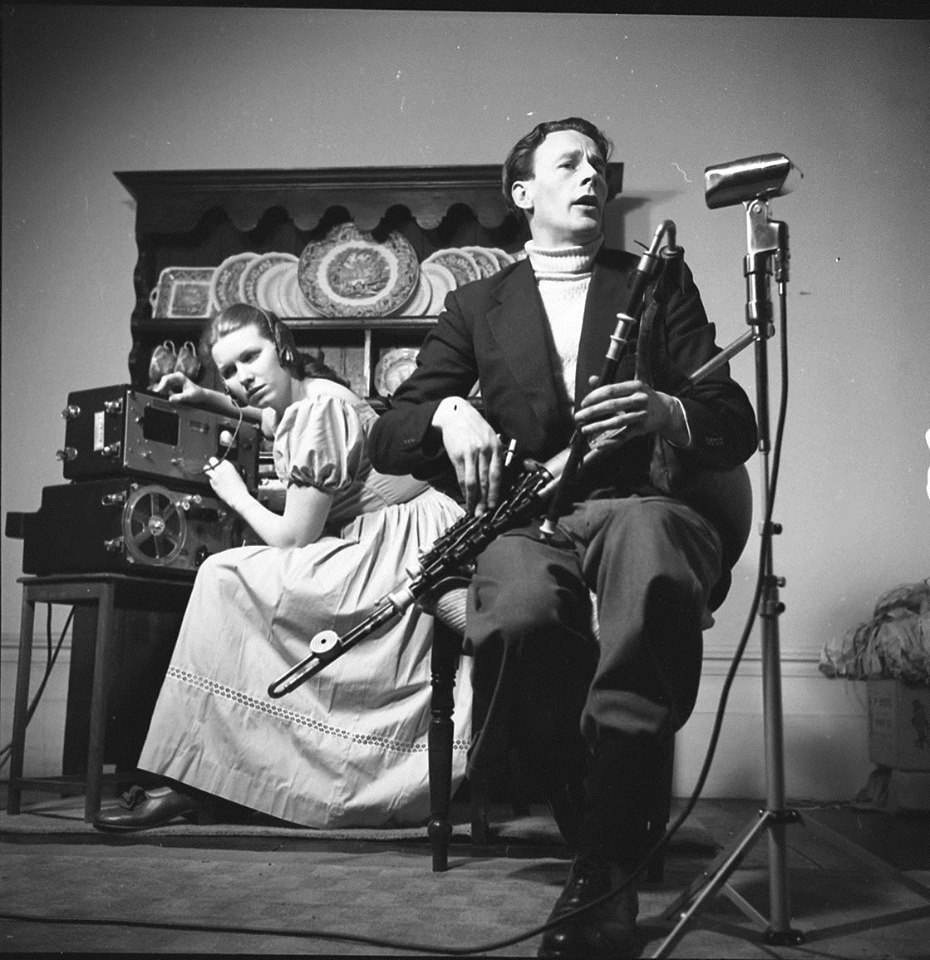 In the foreground, Séamus Ennis sits with his pipes. In the background, Jean Ritchie is leaning intently over her recording equipment.