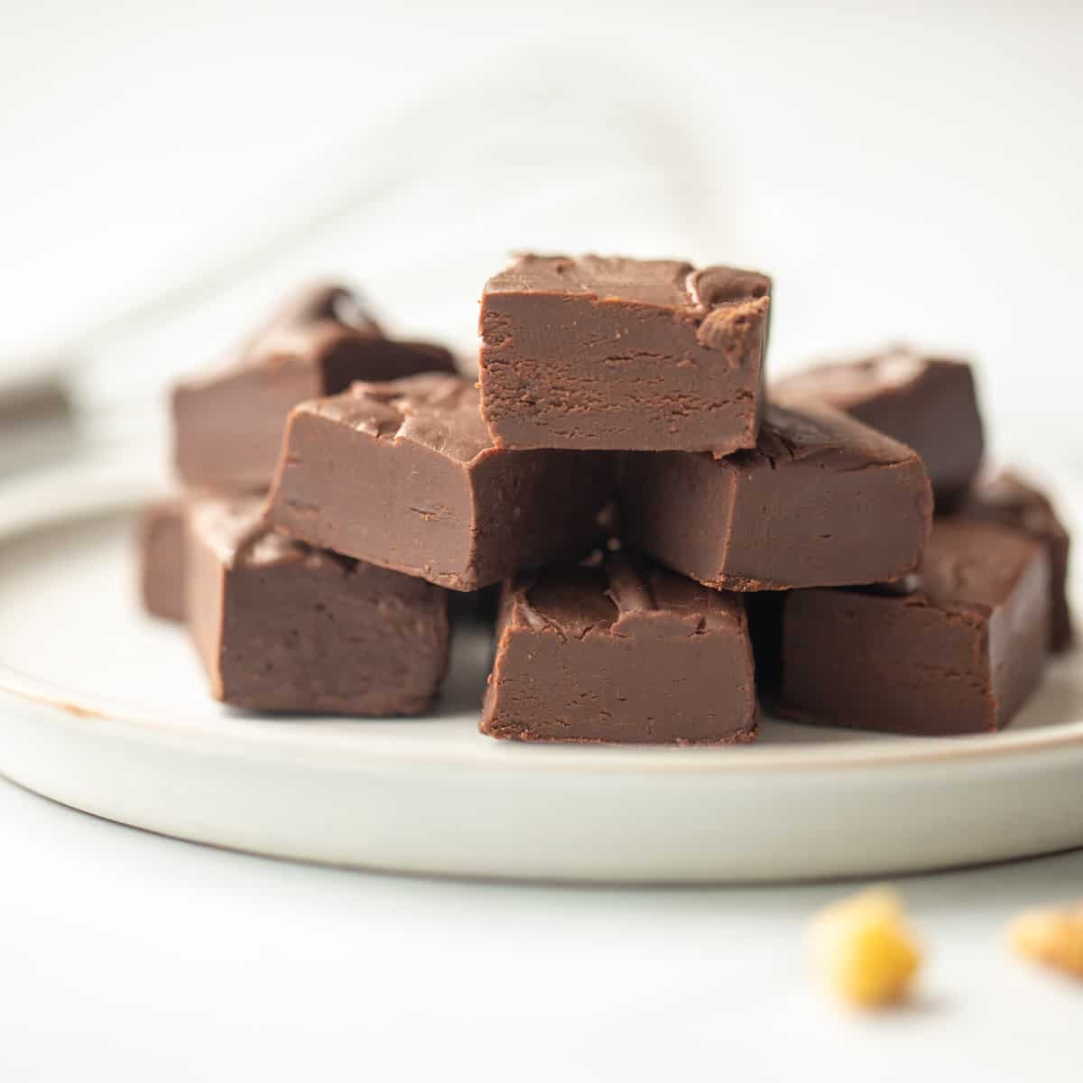 Fudge cut into cubes on white plate with nuts and chocolate chips in background.