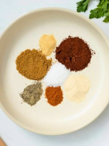 Spices for chili seasoning in cream bowl made with chili seasoning, onion powder, garlic powder, pepper, cumin, salt, and cayenne.