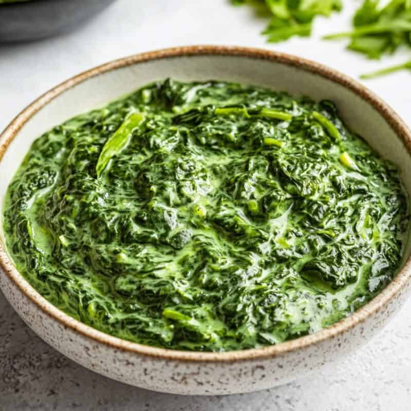 A white ceramic bowl filled with creamy Keto Creamed Spinach on a light stone surface, garnished with a touch of black pepper and fresh spinach visible in the background.