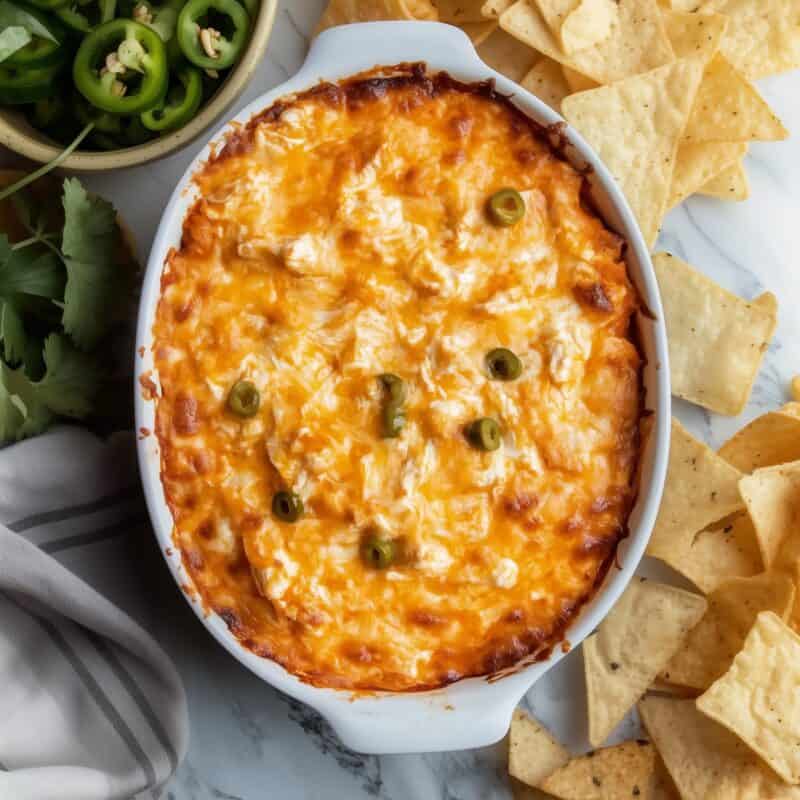 Overhead view of creamy Buffalo Chicken Dip in a white oval dish, garnished with jalapeños and paired with tortilla chips on a marble countertop.