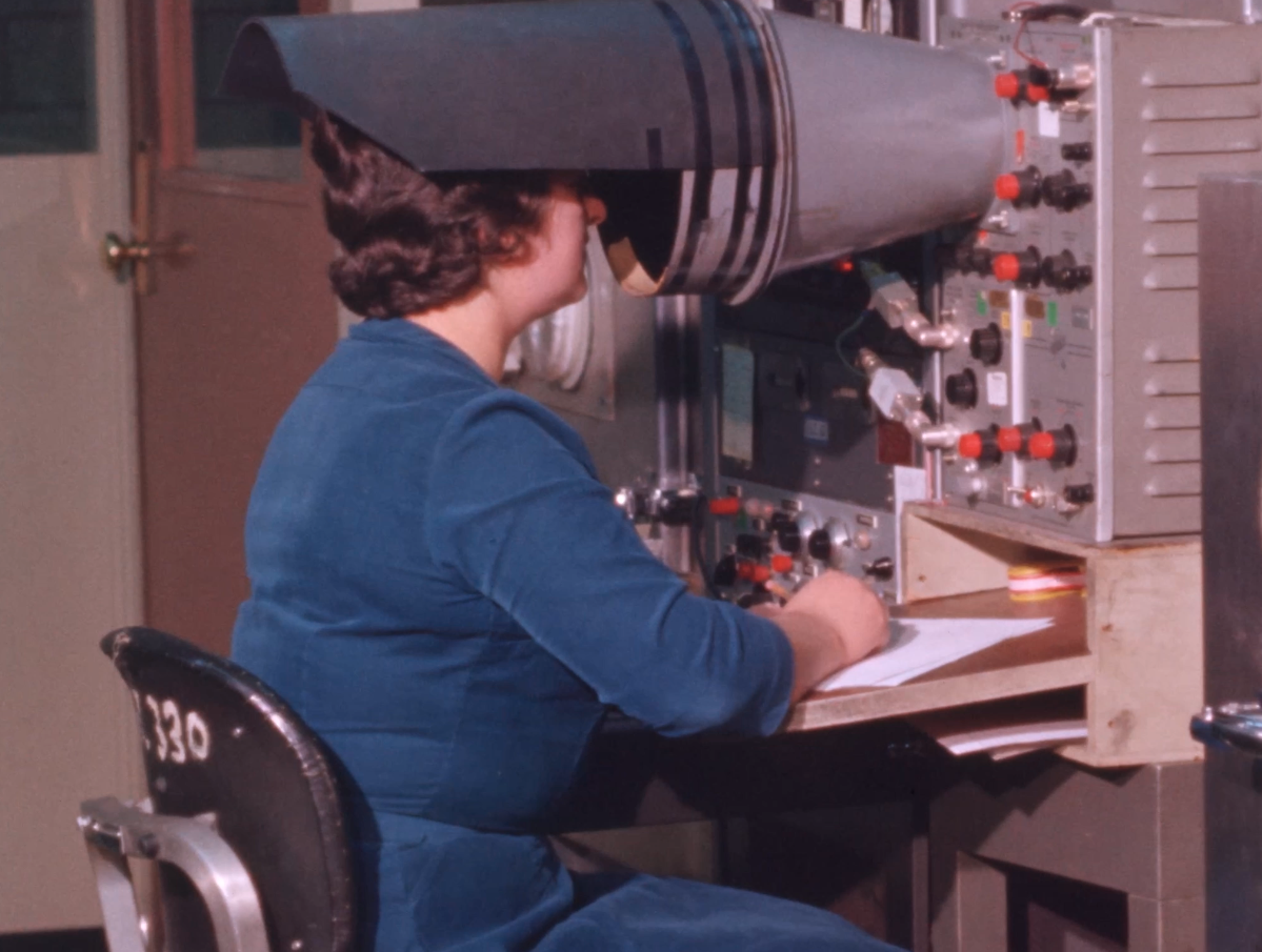 Film still of woman looking into a retro machine with her hands on the control panel.