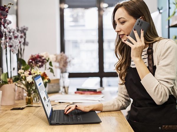 Woman in a shop talking on a cellphone in one hand while her other hand is hovering over a laptop