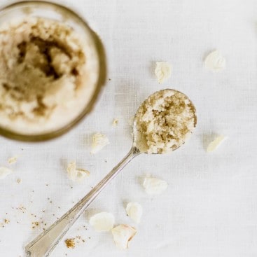 Spoon full of homemade sugar scrub on table with white linen tablecloth.