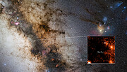 The image shows a section of our galaxy, the Milky Way, in the night sky. This is where the supermassive blackhole and the newly discovered binary star around it are located. The Milky Way traverses the image diagonally and is a spectacle of light and darkness. Its millions of stars illuminate the picture, but light is often blocked by large dark clouds of gas and dust that create eerie shapes. It also contains stars and small nebulae of red, pink and blue colours. In the background, the sky is dark, but full of stars of different sizes and colours. The centre of the galaxy with the binary star appears in a close-up on one corner of the image. This is a pixelated mapping of the hydrogen emission of the region around the star pair D9. Bright orange to white patches represent high hydrogen levels (indicating stars), black, represents very low hydrogen emission. Most of the centre of the image is dark with a few wisps of dark orange. The black hole and the binary D9 are indicated.