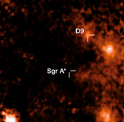 The image is a pixelated mapping of the hydrogen emission of the region around the star pair D9. Bright orange to white patches represent high hydrogen levels (indicating stars), black, represents very low hydrogen emission. Most of the centre of the image is dark with a few wisps of dark orange. The location of the supermassive black hole Sagittarius A* is indicated by two lines in the middle of the image, in a black patch. Two lines indicate the location of the star pair D9, which is showing as a bright orange patch. Most of the image is dark, with three bright orange-reddish patches at the right, and one at the bottom left.