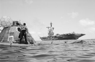 Command module floating on the water and a large Navy ship in the background. A recovery crew member is pictured standing on the command module