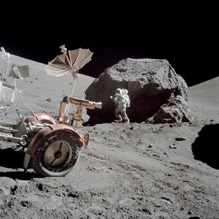 Harrison H. Schmitt collects samples from a huge boulder that is much taller than him. Part of the lunar roving vehicle is visible in the foreground.