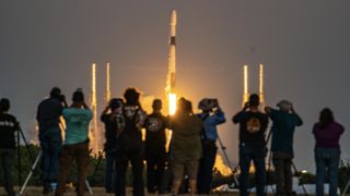 a row of people in the distance take photos of a rocket far away but large in frame.