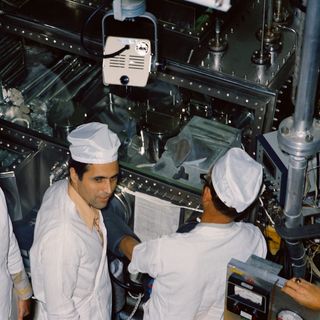 Scientist-astronaut Harrison H. Schmitt faces the camera wearing a white protective suit and white hat. Another man is standing next to him with his back to the camera.