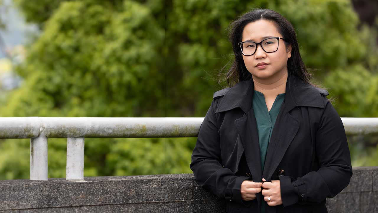 A young asian woman with dark hair looks at us seriously, hands clutching her black jacket. She stands on a bridge, foliage behind her.