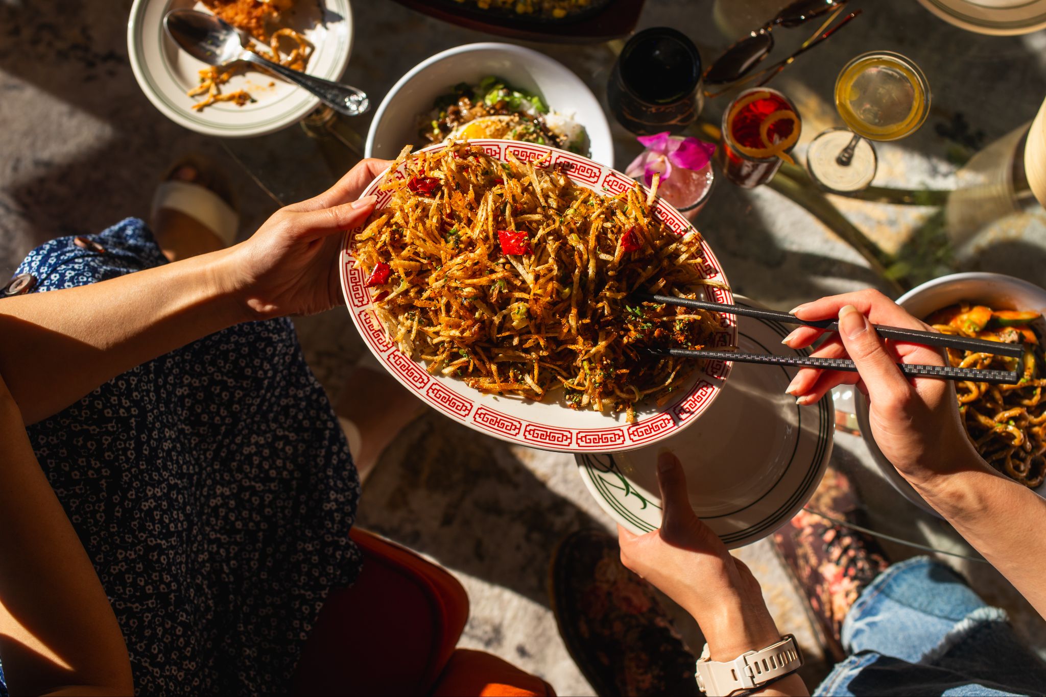 Two pairs of outstretched hands passing a plate piled with pan-fried noodles over a sun-dappled table.