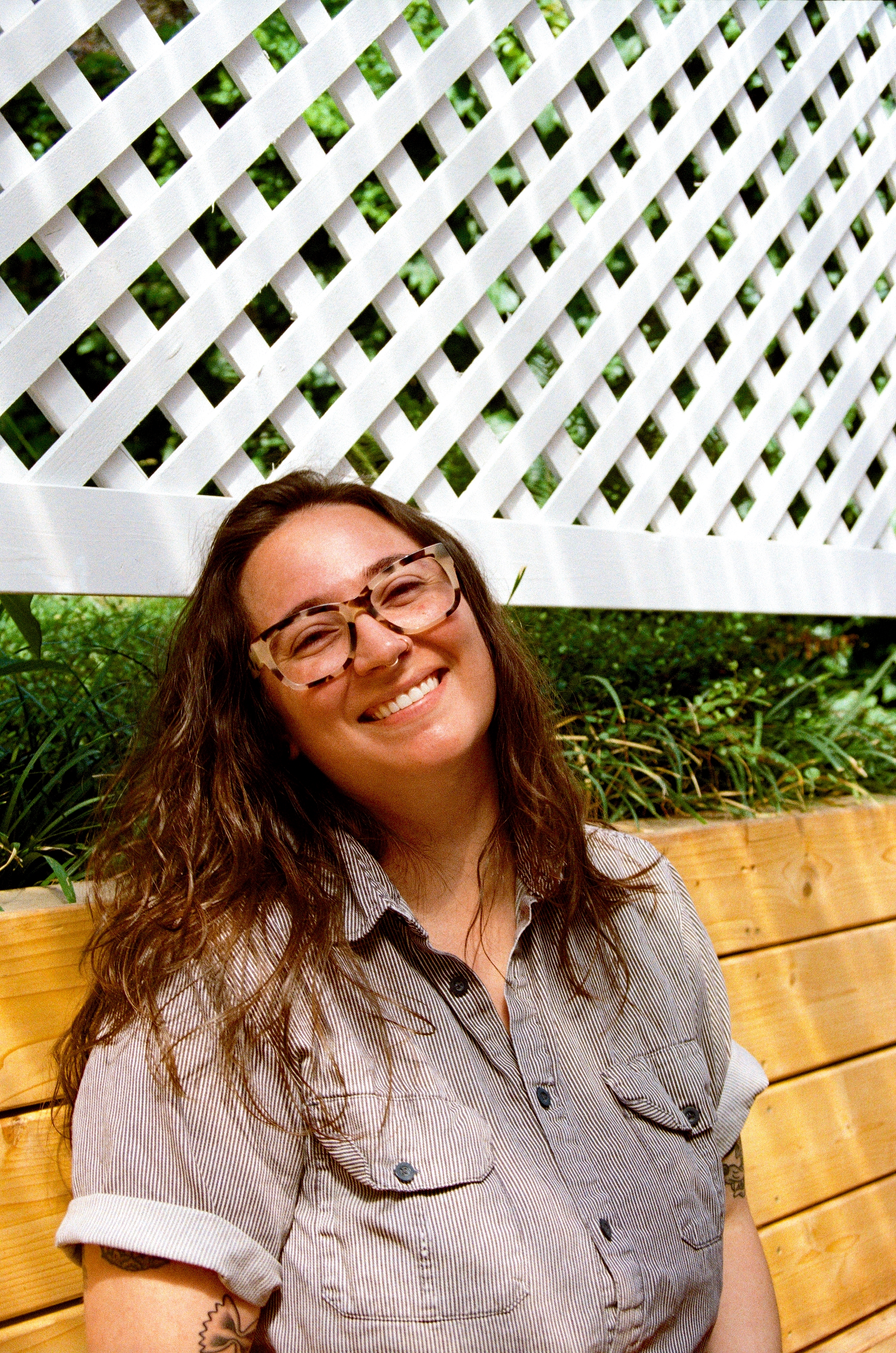 A smiling woman wearing glasses and a collared short-sleeve shirt stands in front of a makeshift white fence.