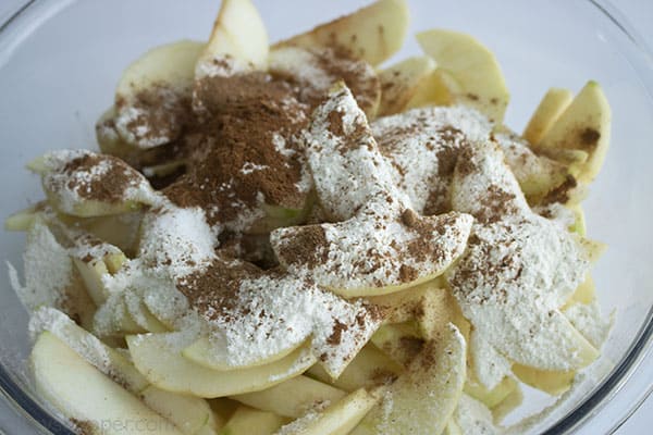 apple slices in a large clear bowl with flour, sugar, and cinnamon on top 