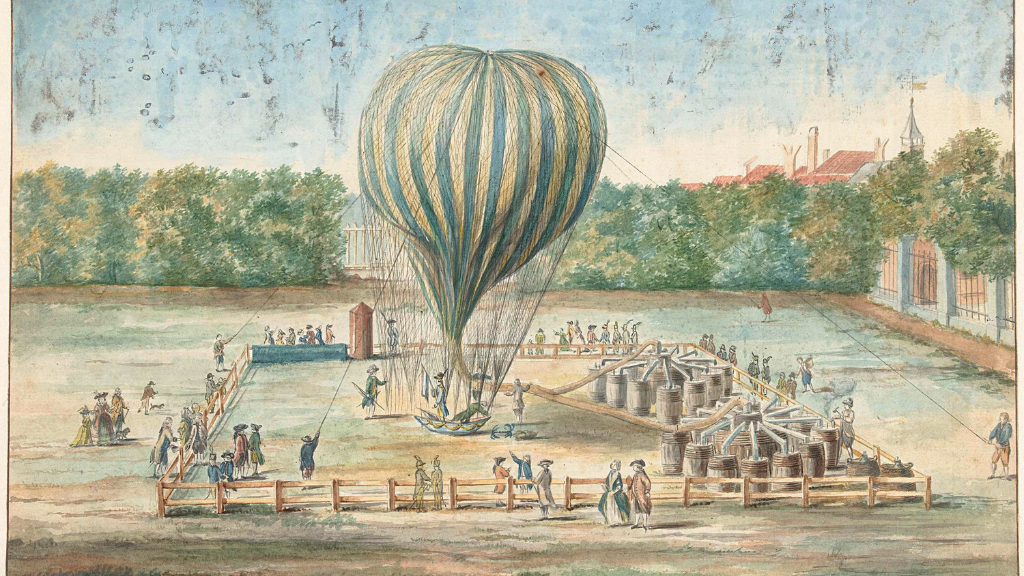 Fancily-dressed people gathered in a field surrounded by trees and a tall fence for the launch of a blue-yellow striped hot-air balloon held by long strings.