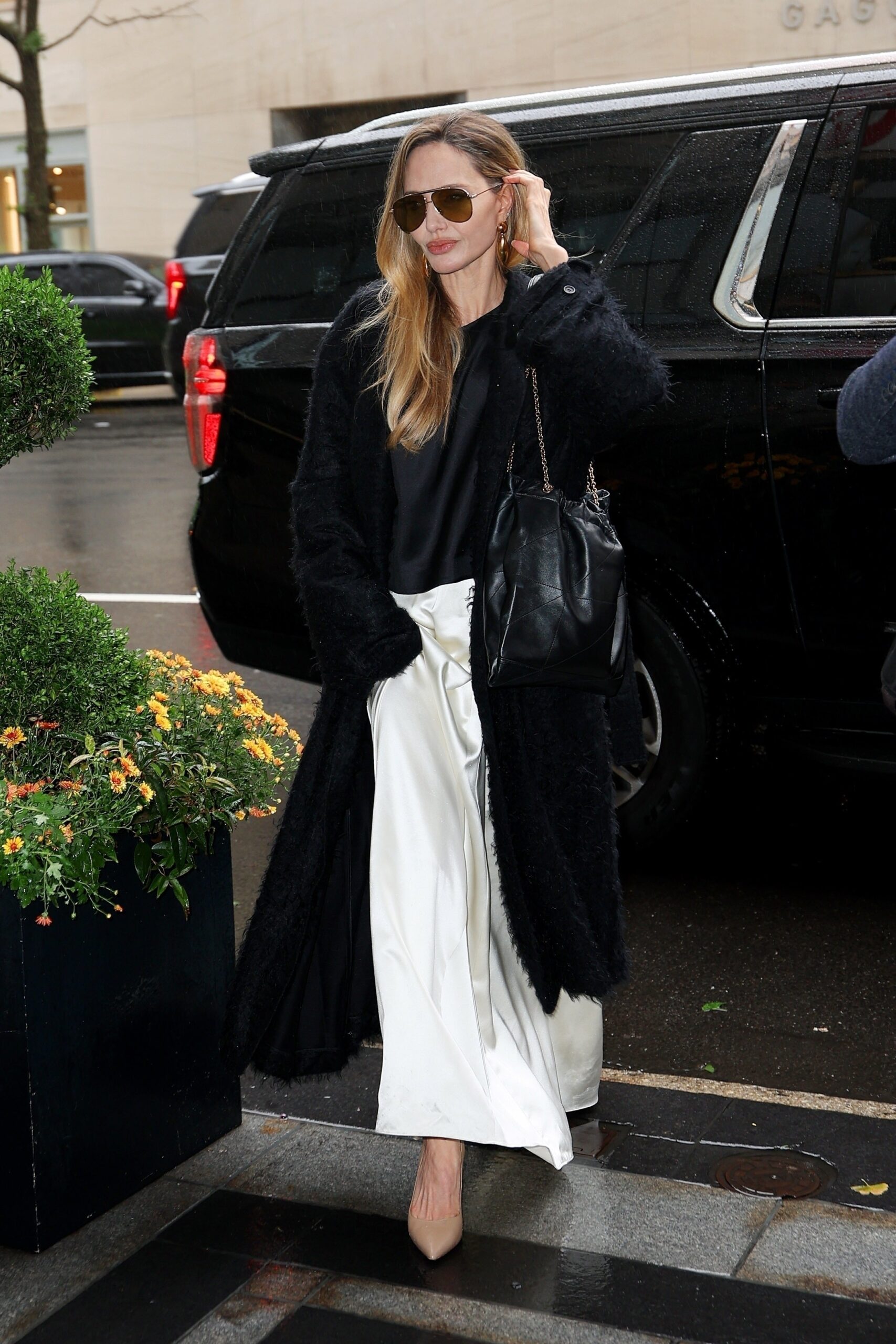 CR25 cover star Angelina Jolie radiated elegance on a rainy overcast day in New York City in a black and white ensemble styled with a Saint Laurent by Anthony Vaccarello handbag. The actress stepped out of her car in delicate pointed toe nude heels, a creamy silk skirt, black silk top, and a slightly oversized textured black coat. The Saint Laurent Jamie 4.3 Pochon bag in lambskin gave a touch of luxurious practicality to Jolie’s look. The bag’s gold chain handles complimented Jolie’s chunky gold earrings and warm tinted aviator sunglasses. Jolie’s blonde hair in natural waves was the final touch to her effortlessly chic rainy day look.  Courtesy of Saint Laurent