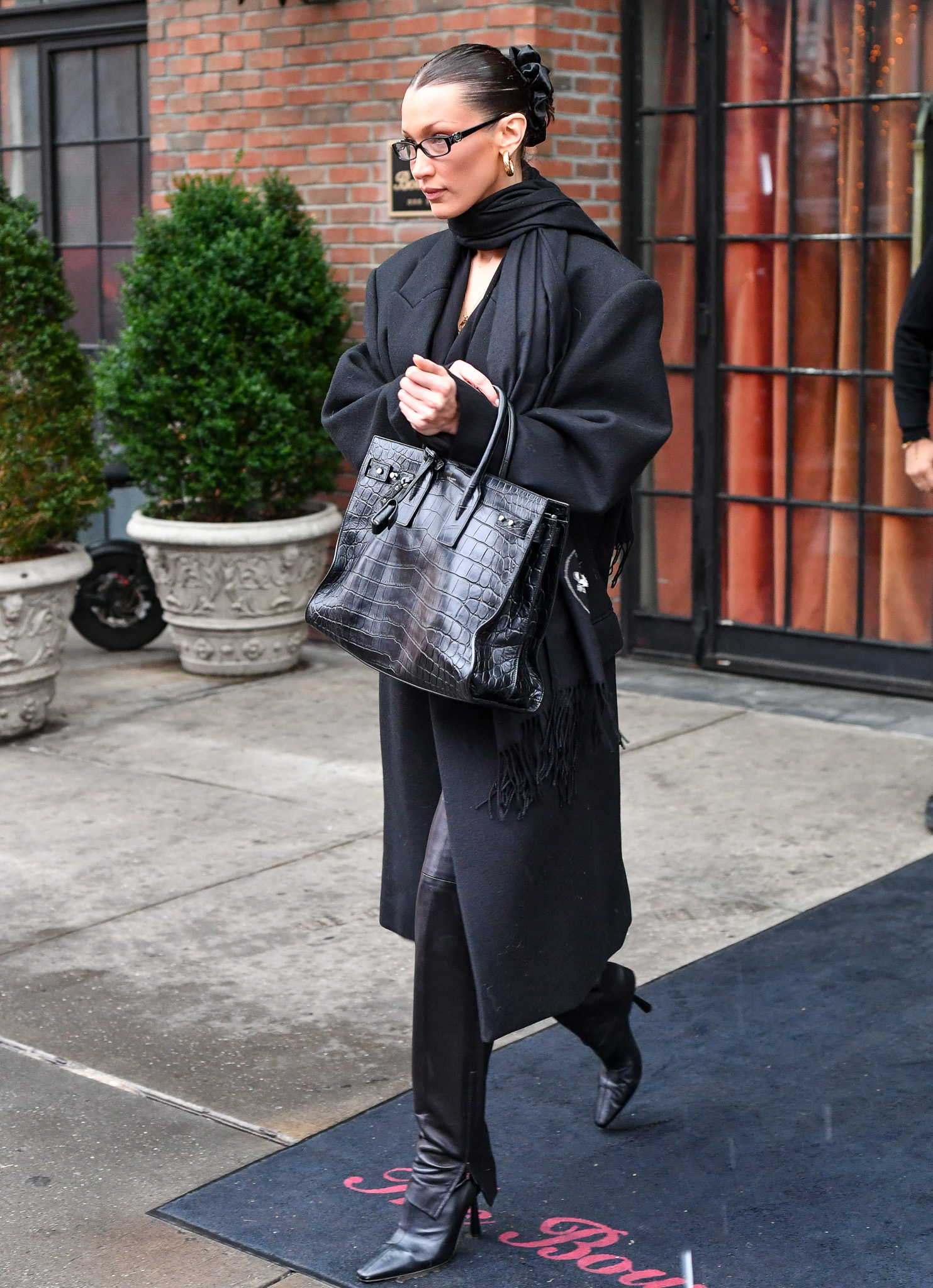 Bella Hadid was spotted on the streets of New York City wearing a luxurious and monochromatic look. The supermodel sensation looked impossibly chic in a long black coat, black leather pants, and pointed black boots. Hadid styled her winter weather ensemble with a black scarf draped over her shoulder, narrow frame glasses, oversized hoop earrings, and her signature slick bun. However, the star of Hadid’s all-black look was the Saint Laurent by Anthony Vaccarello Sac de Jour handbag in crocodile-embossed leather, completing the model’s chic city outing.  Courtesy of Saint Laurent