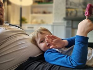 Boy lying on Dan, waving, eating an ice lolly.