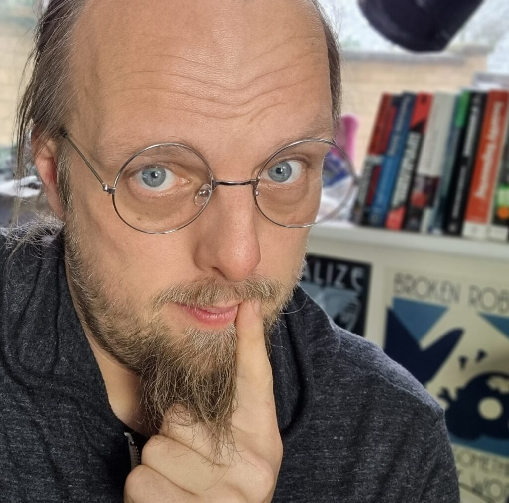 A white man wearing simple round spectacles with a silver wire rim, his finger to the corner of his mouth, looks into the camera.