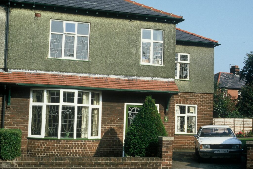 A partially-pebbledashed house, number 7, with an old white Ford Escort parked in the driveway.