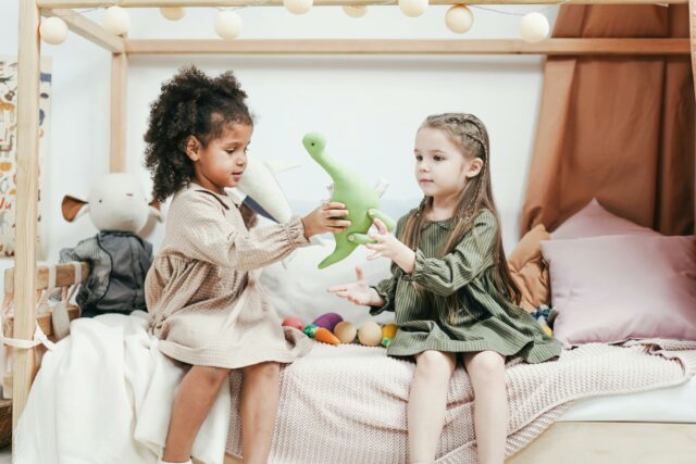 Two children sit on a bed: one hands a toy dinosaur to the other.