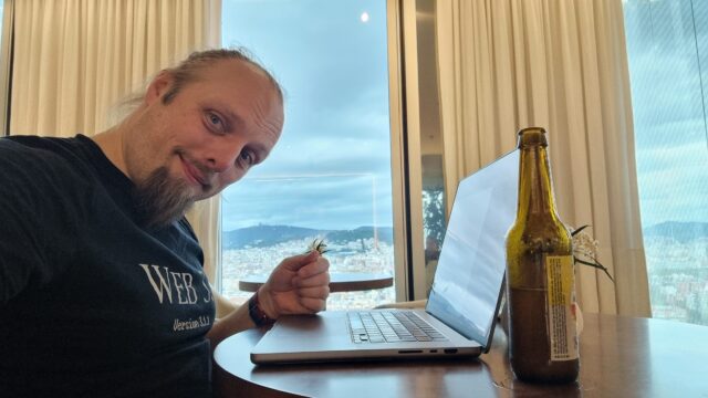 Dan sits at a laptop in a hotel bar, a view of Barcelona out of the window behind him, a beer bottle alongside him.