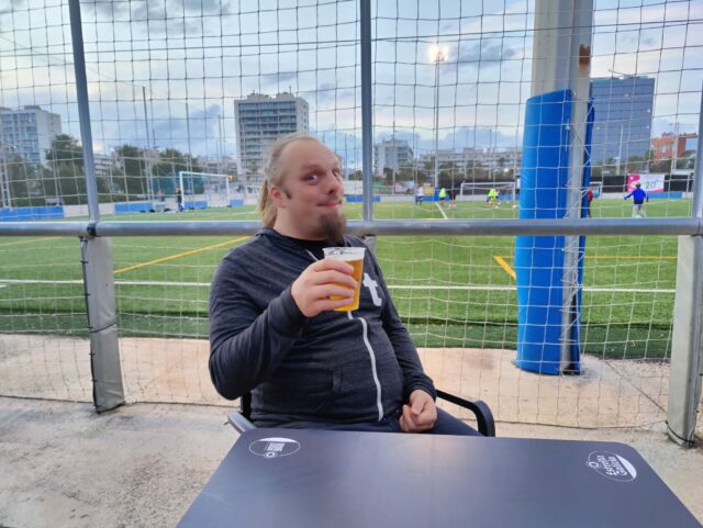 Dan drinks a beer, a children's football match is being played in the background.