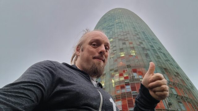 Under a cloudy early-morning sky, Dan gives a thumbs-up in front of a gherkin-shaped skyscraper.