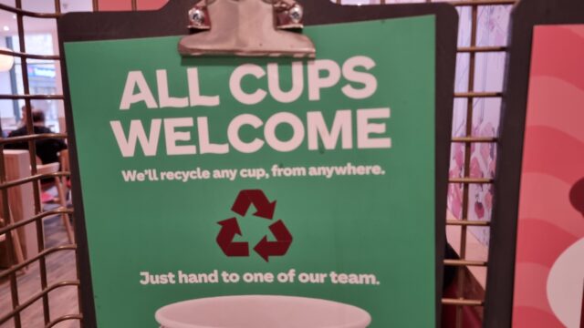 Sign made from a clipboard, in a coffee shop, reading "All cups welcome. We'll recycle any cup, from anywhere. Just hand to one of our team."