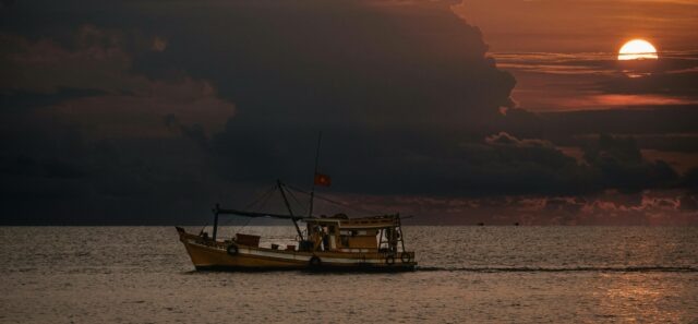 As the sun sets behind growing clouds, a small fishing vessel flying a red flag glides across a moderately-smooth ocean.