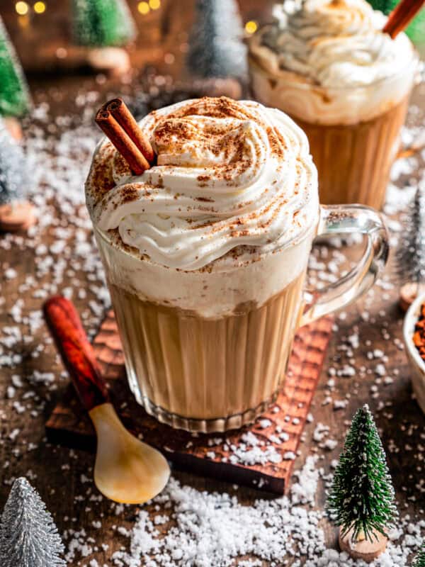 A Starbucks eggnog latte garnished with whipped cream, ground nutmeg, and a cinnamon stick in a glass mug, with second mug of latte in the back.