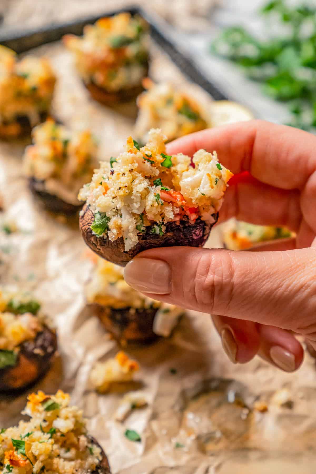 A hand holding up a crab stuffed mushroom with more stuffed mushrooms on a baking sheet in the background.