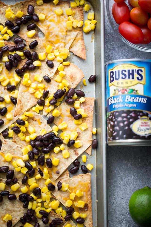 Overhead view of canned corn and black beans layered over tortilla chips on a sheet pan.