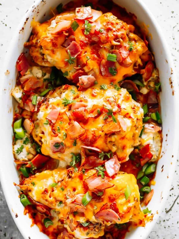 three baked chicken breasts in a white baking dish, and topped with melted cheese, crumbled bacon, and sliced green onions.